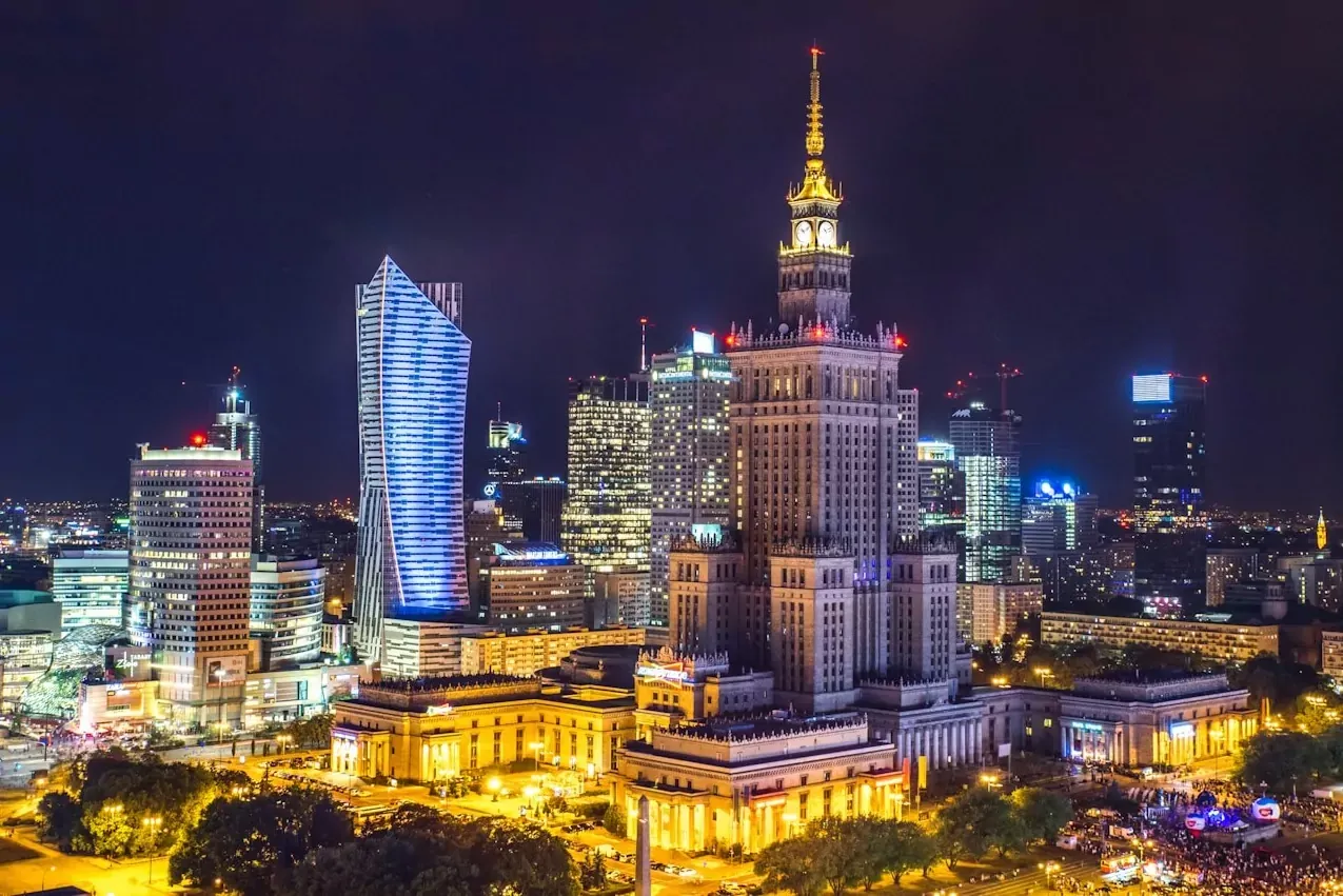 Warsaw city skyline at night featuring the illuminated Palace of Culture and Science, modern skyscrapers, and city lights.