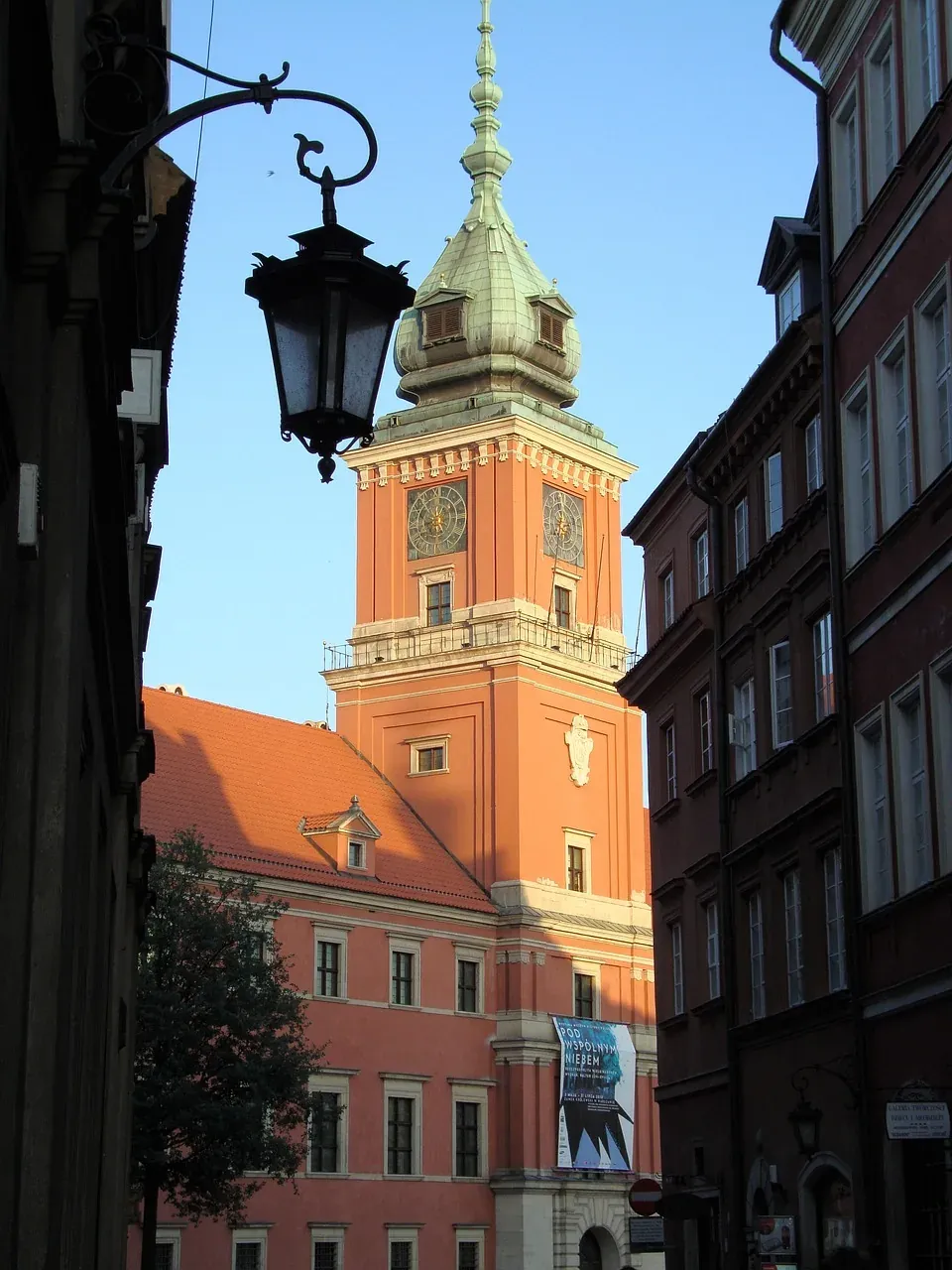 A peach-colored clock tower with a green dome and two clock faces, framed by narrow historic buildings and a black ornate str