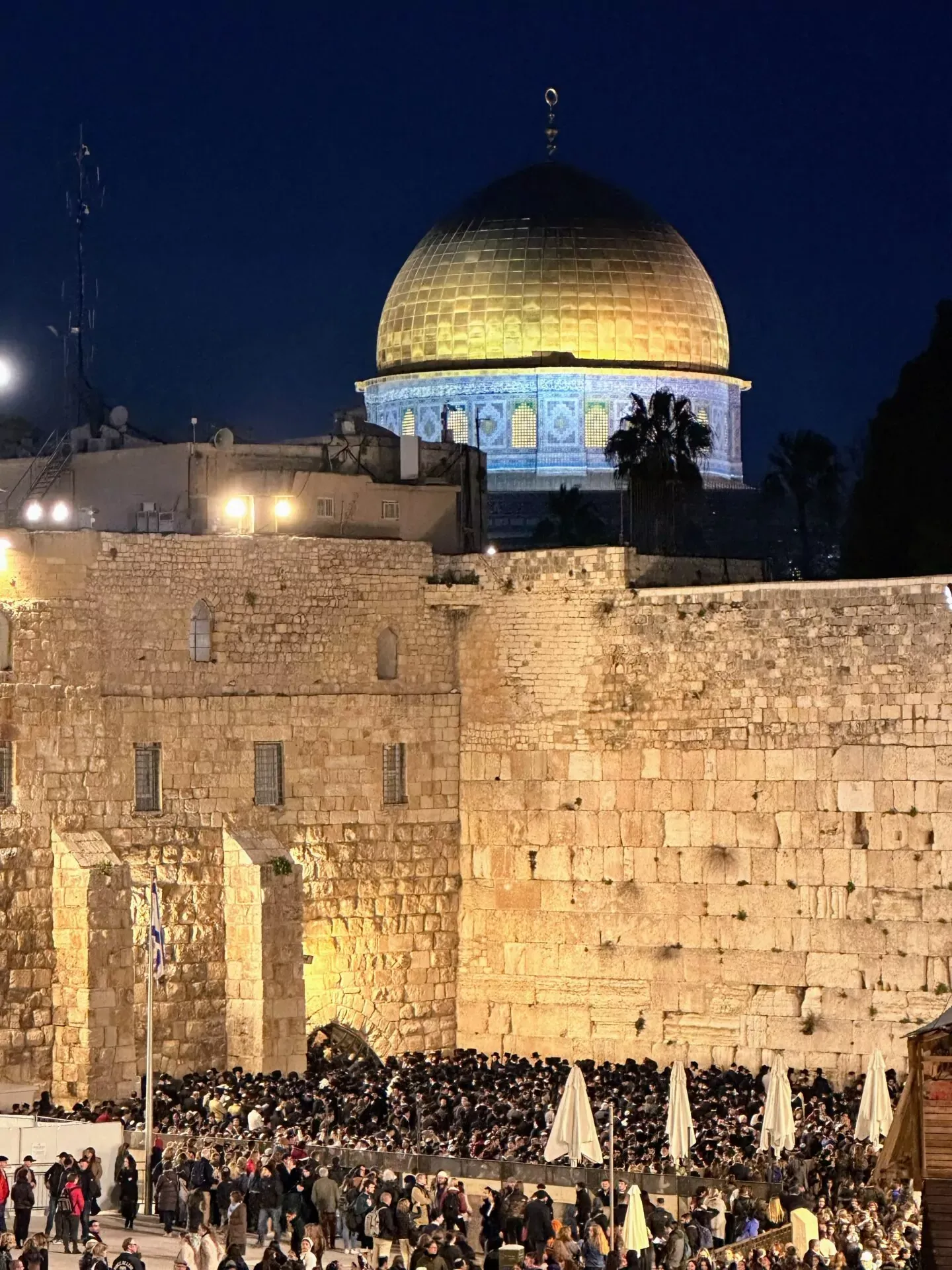 The Dome of the Rock with its golden dome illuminated at night, towering above the stone Western Wall and a large crowd.