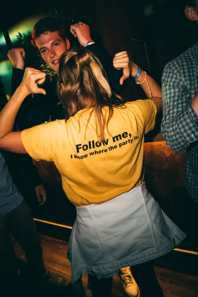 People dancing in a dimly lit bar, one person in a yellow t-shirt with 'Follow me, I know where the party is' on the back.