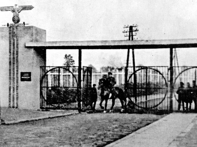Black and white photo of a camp entrance gate with a Nazi eagle emblem, men in uniform, and circular metalwork.