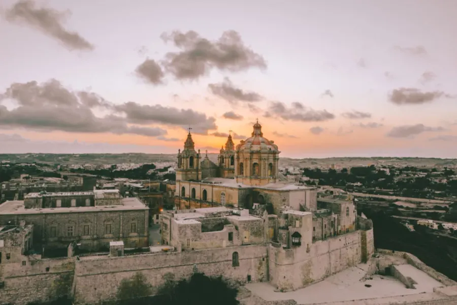 Aerial view of Mdina's St. Paul's Cathedral with domes and bell towers, surrounded by fortified walls and historic buildin...
