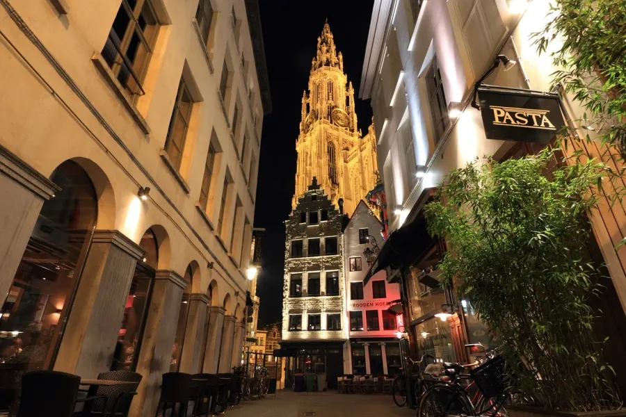 Illuminated Antwerp Cathedral at night, seen from a charming street with restaurants.