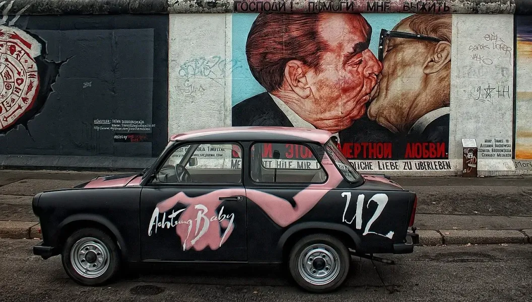 A Trabant car with graffiti art parked near the iconic Brotherly Kiss mural on the East Side Gallery in Berlin.