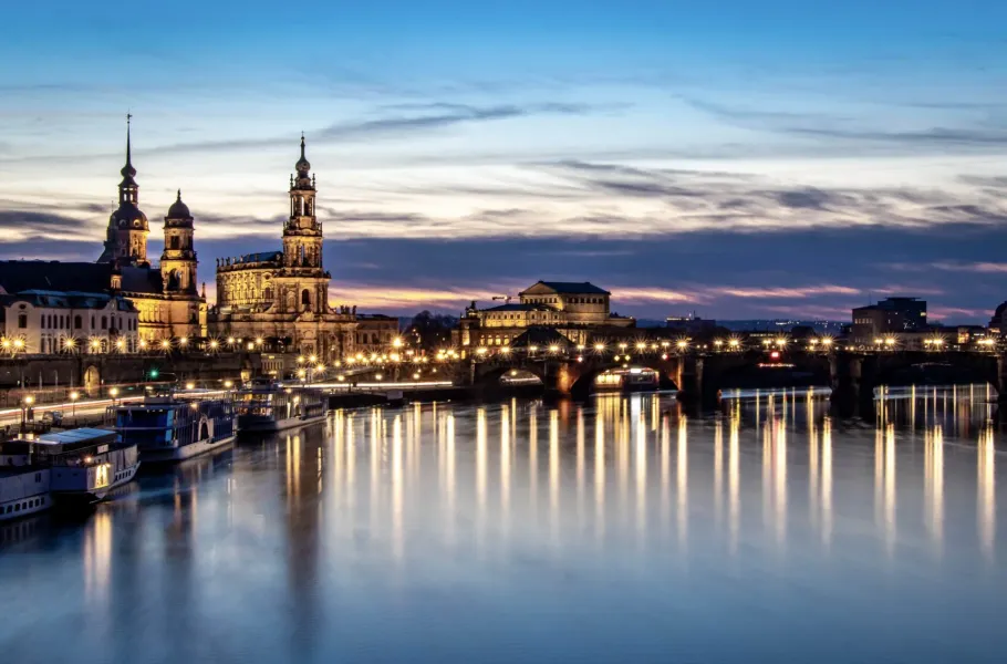 Illuminated Dresden skyline at dusk with Frauenkirche, Hofkirche, Augustusbrücke bridge, and boats on the calm Elbe River.