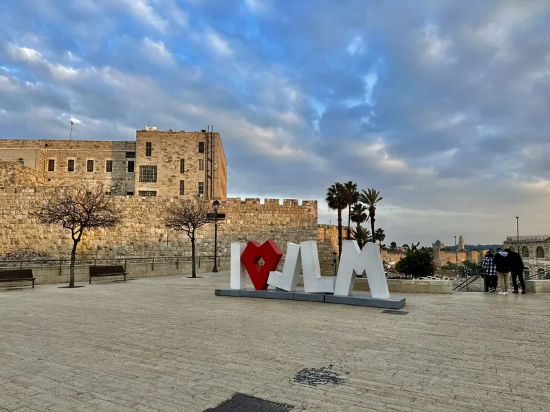 Tourists visit the iconic "I ♥ JLM" sign in Jerusalem, with the Old City walls in the background.