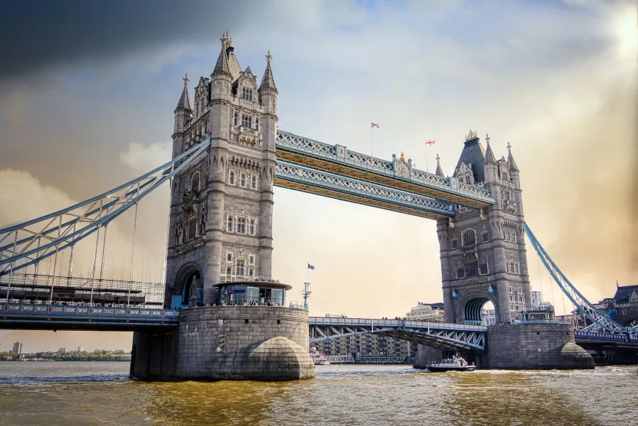 Tower Bridge spanning a wide river under a partly cloudy sky, with a boat on the water and buildings in the distance.