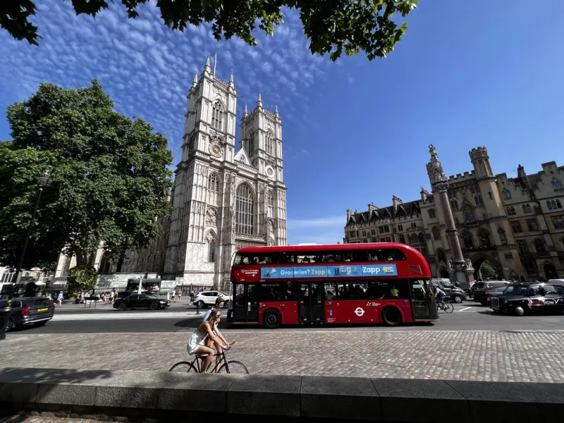 Westminster Abbey's historic facade and tower under a blue sky, with a red double-decker bus, cars, and people on the street.