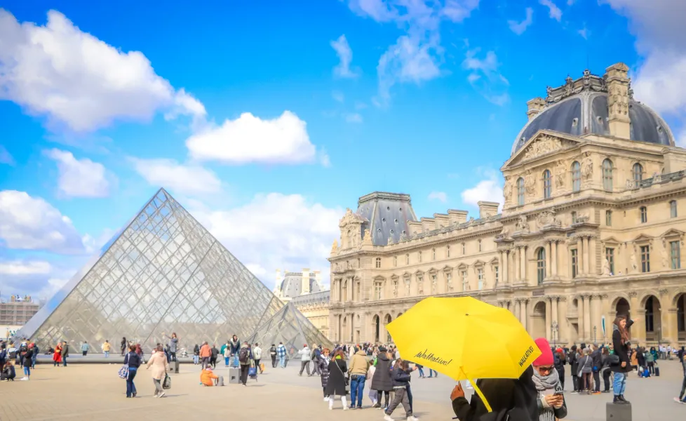 The glass pyramid entrance of the Louvre Museum on a sunny day with clouds, surrounded by classical palace buildings and v...
