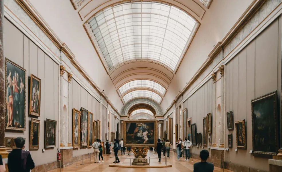 Tourists exploring a grand gallery at the Louvre Museum in Paris, surrounded by classical paintings.
