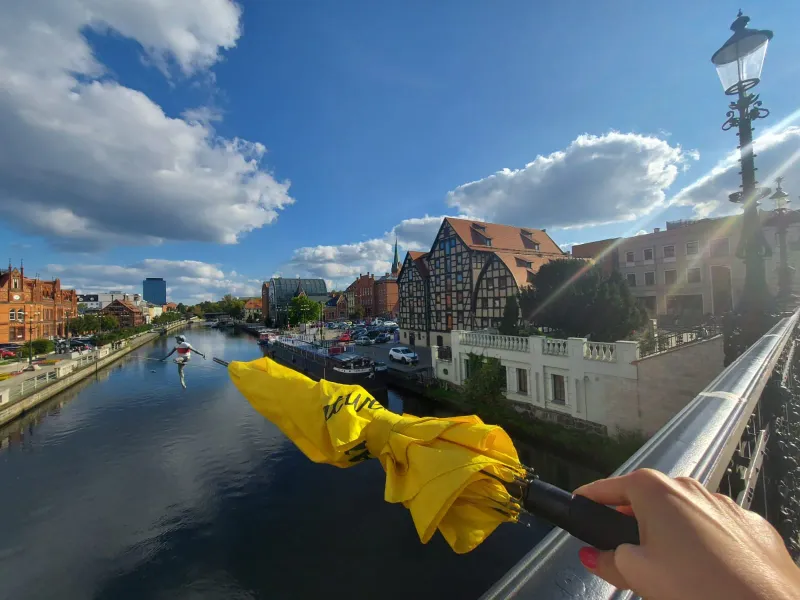 A yellow umbrella held on a bridge overlooking a river with historic buildings and a tightrope walker statue.