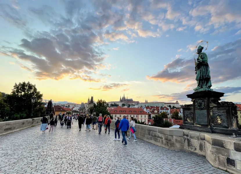Tourists walking across Charles Bridge in Prague at sunset, with Prague Castle in the background.