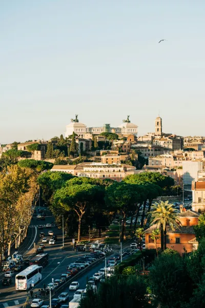 Panoramic view of Rome with the Victor Emmanuel II Monument atop a hill, a busy street with cars and buses, and green trees.