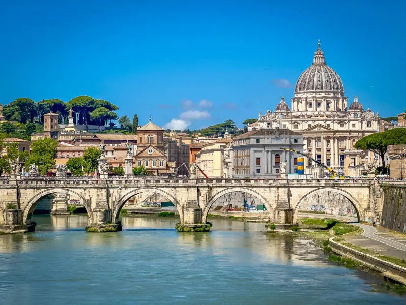 Sant'Angelo Bridge over the Tiber River, with St. Peter's Basilica dome and Roman buildings under a clear blue sky.