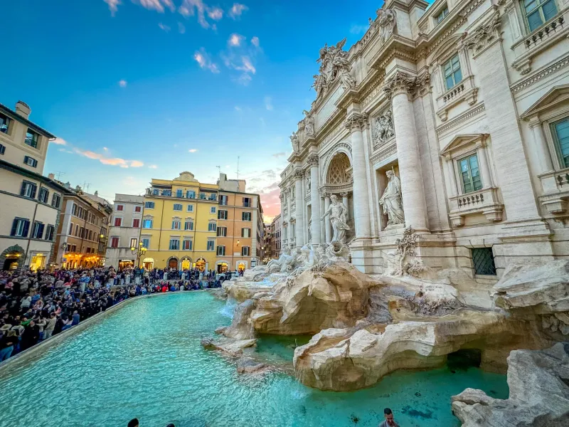Ornate white Trevi Fountain in Rome with turquoise water, surrounded by a large crowd of people under a blue and pink sky.