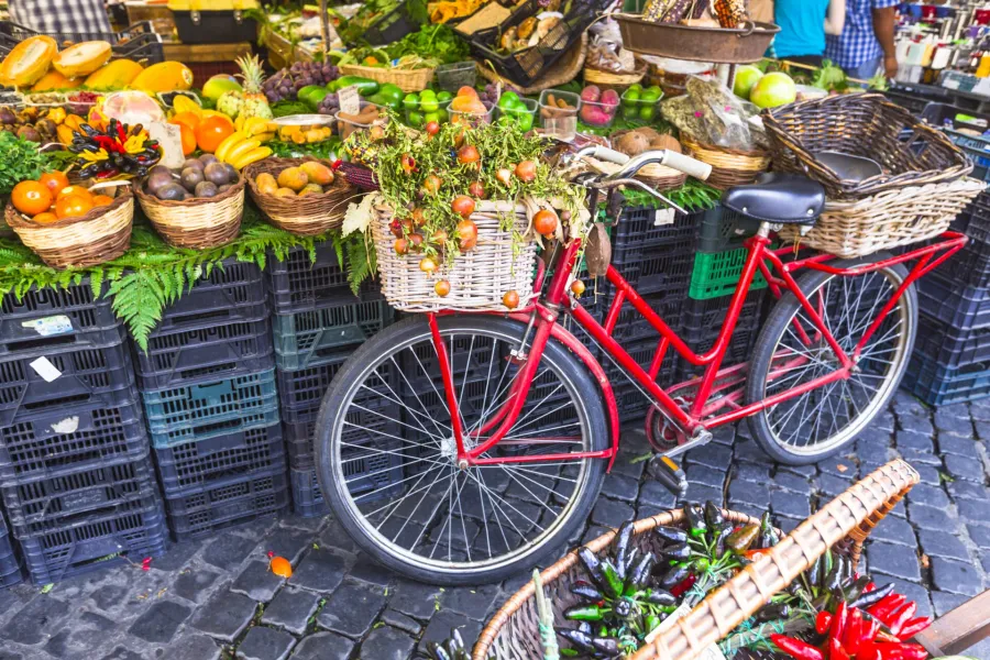 Red bicycle with fruit in basket at a vibrant Roman market.