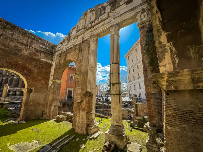 Ancient Roman brick ruins with arches and two tall stone columns. Green grass fills the excavated area, modern buildings v...