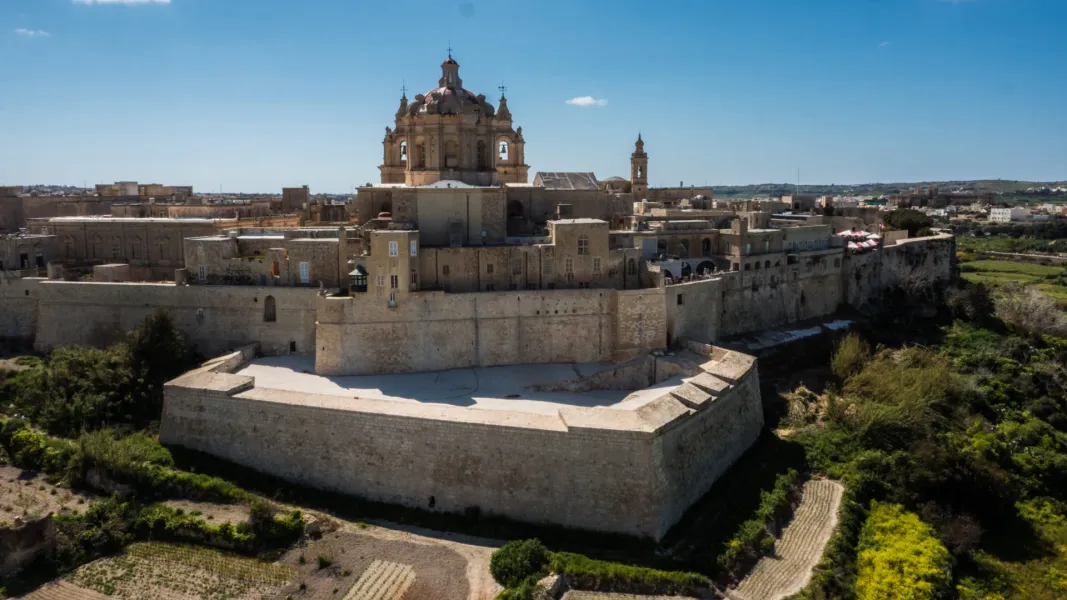 Aerial view of a historic fortified city with golden stone walls, buildings, and a large domed cathedral under a blue sky.
