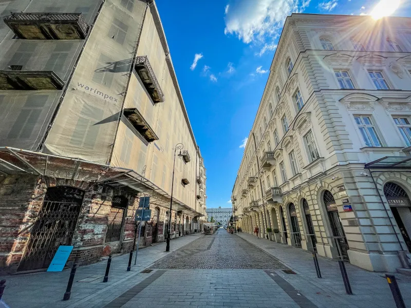 Sunlit cobblestone street in Warsaw, Poland, with historical buildings and one under renovation.