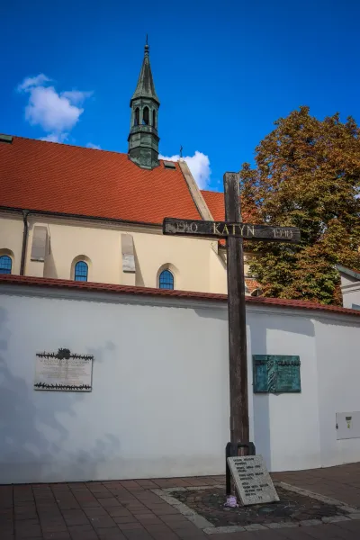 Large wooden Katyn Cross memorial with 'KATYN 1940 1990' inscription, plaques on a white wall, church roof, and spire.