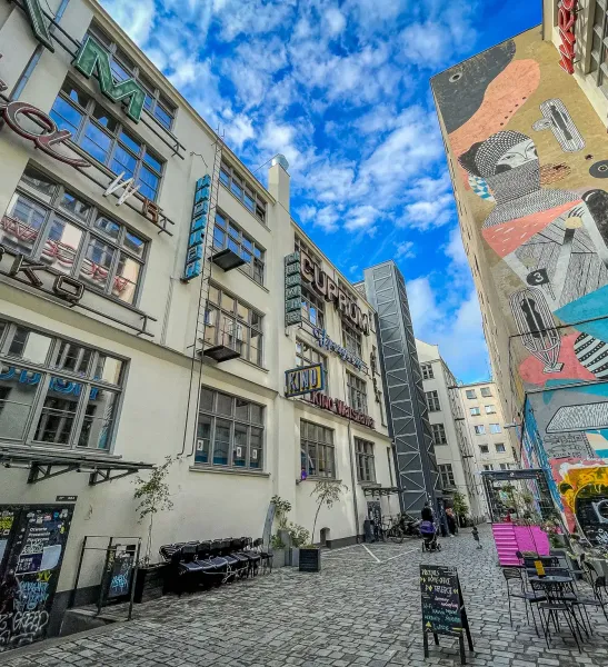 A narrow cobbled alleyway in Wrocław, flanked by a building with neon signs and another with a large abstract mural. Outdo...