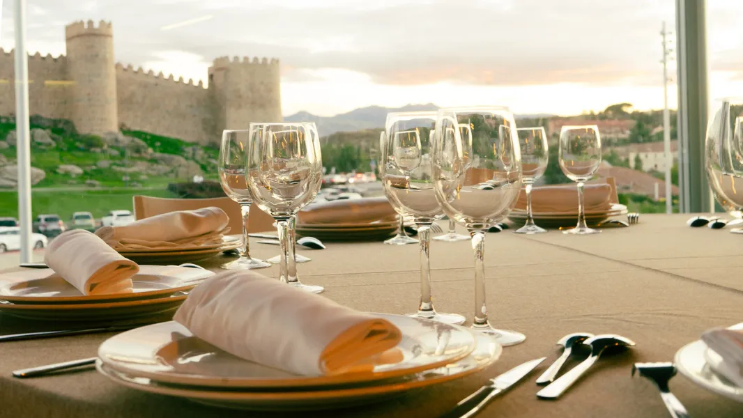 A dining table set with glasses and plates, overlooking the medieval city walls of Ávila, Spain at sunset.