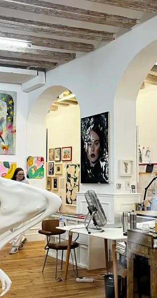Art studio interior with paintings on white walls, exposed wooden beams, a partial white sculpture, and a woman observing ...