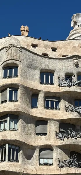 Close-up of Casa Milà's wavy stone facade with dark windows, grey roller blinds, and wrought-iron balconies under a blue sky.