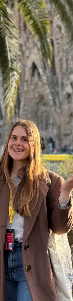 Smiling woman with long brown hair, brown coat, and yellow 'Catalonia Tourism Guide' lanyard. Blurry building and palms in...