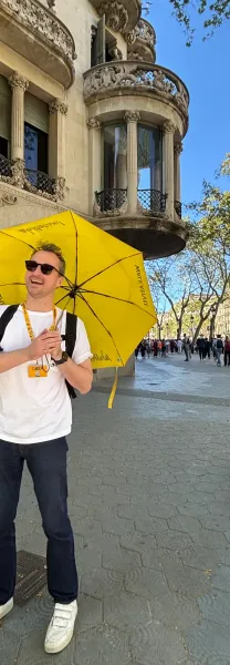 A smiling male tour guide holding a yellow Walkative umbrella on a sunny street in Barcelona. Ornate modernist building in...