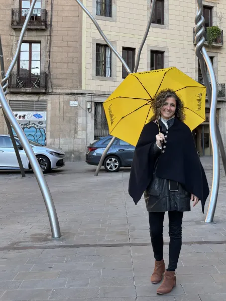 Smiling woman with curly hair holds a yellow Walkative umbrella in Plaça de Sant Miquel, Barcelona. Buildings and cars beh...