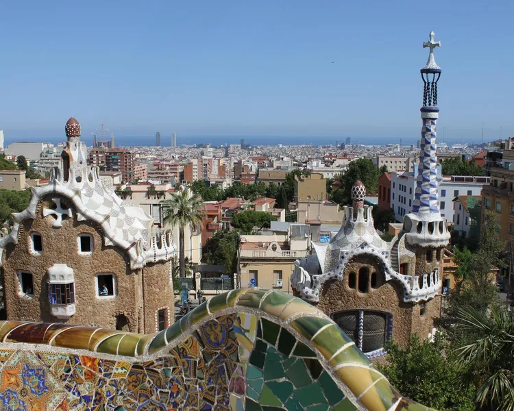 View from Park Güell showing two Gaudi-designed mosaic-roofed buildings, a colorful tiled wall, and Barcelona cityscape wi...