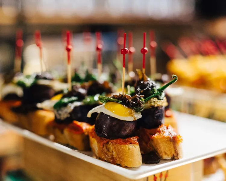 Close-up of a plate of pintxos, small open-faced sandwiches with various toppings, held by red toothpicks. A dark sausage ...