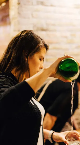 Person pouring sidra from a green bottle into a glass, generating foam. Blurred stone wall in background.