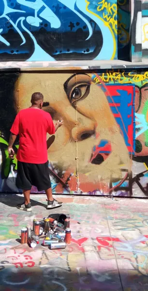 A person in a red t-shirt spray painting a large mural of a woman's face on a wall covered in graffiti. Spray cans on the ...