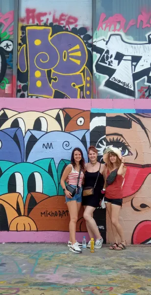 Three women smiling in front of a colorful wall covered in abstract graffiti and a large mural with stylized faces.