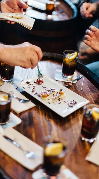 People's hands around a wooden table with tapas plates and drinks. One person uses a fork to eat a dish.