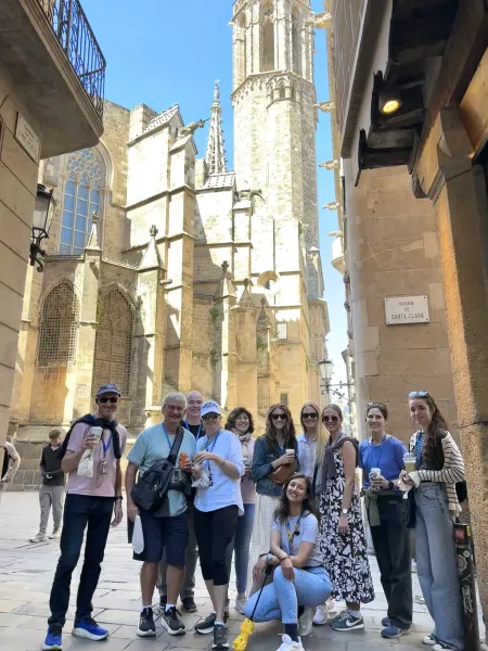A diverse group of people stands on a narrow stone street with Barcelona Cathedral in the background under a blue sky.