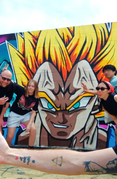 Four people posing in front of a colorful graffiti mural of a character with spiky yellow hair and blue eyes, under a clea...