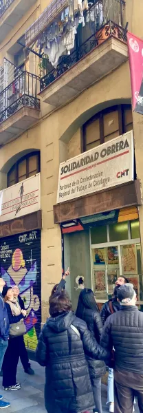 A group of people on a street tour looking up at a building with "Solidaridad Obrera" and "CNT" signs, and graffiti on a s...