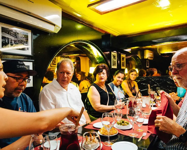 A group of people sitting at a restaurant table with red tablecloths, sharing tapas dishes, olives, and a pitcher of red s...