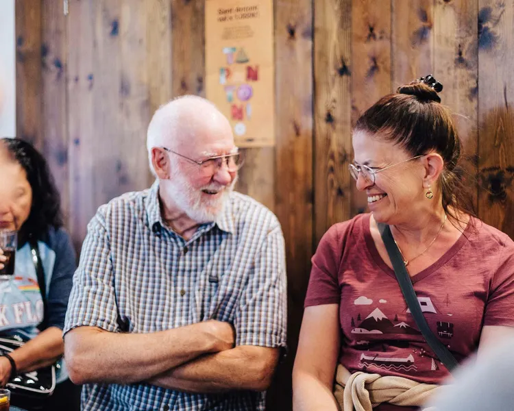 Two smiling travelers, an older man and a woman, talking in a casual setting with a wooden wall and a tapas poster.