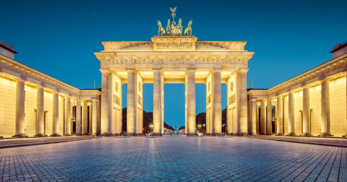The Brandenburg Gate in Berlin, illuminated at dusk, featuring the Quadriga sculpture on top and a wide, empty cobblestone...