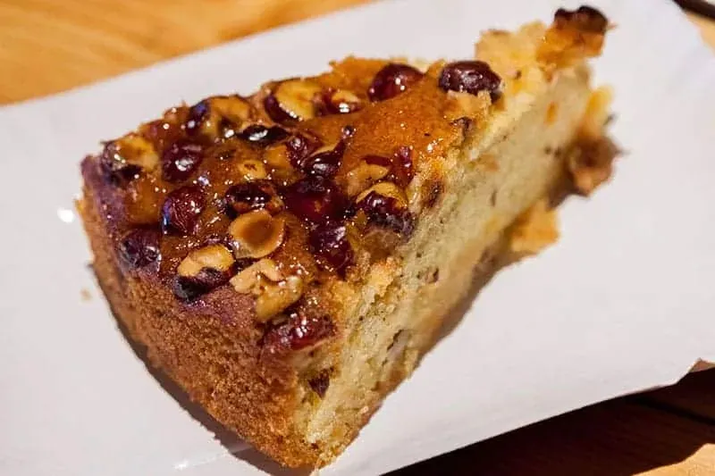 Slice of cake with a glossy topping of roasted hazelnuts and dried berries on a white plate.
