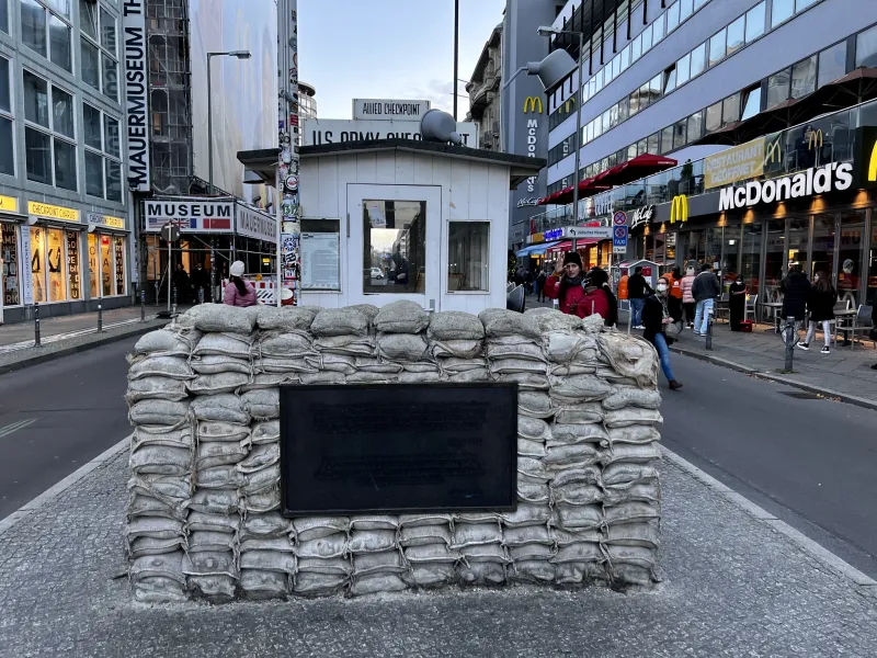Reconstruction of Checkpoint Charlie guardhouse with sandbags and a plaque on a cobblestone street in Berlin, with buildin...