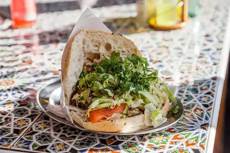 Half a doner kebab in pita bread with meat, lettuce, parsley, and tomato, served on a metal plate on a tiled table.