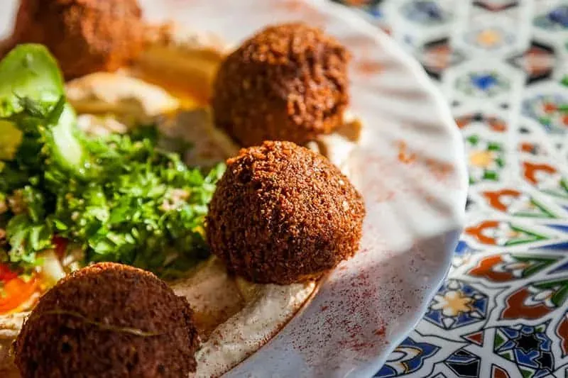 Close-up of a plate with several golden-brown falafel balls, a spread of hummus, and green chopped parsley salad.