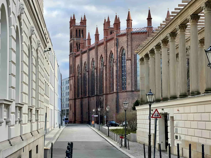 The red-brick Friedrichswerder Church seen from a quiet side street next to a building with a classical colonnade in Berlin.