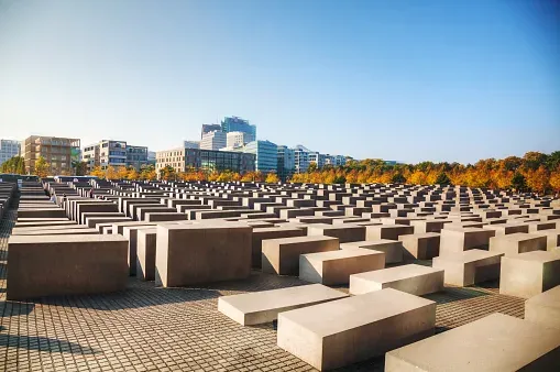 Concrete stelae of the Memorial to the Murdered Jews of Europe in Berlin on a sunny day with city buildings in the backgro...