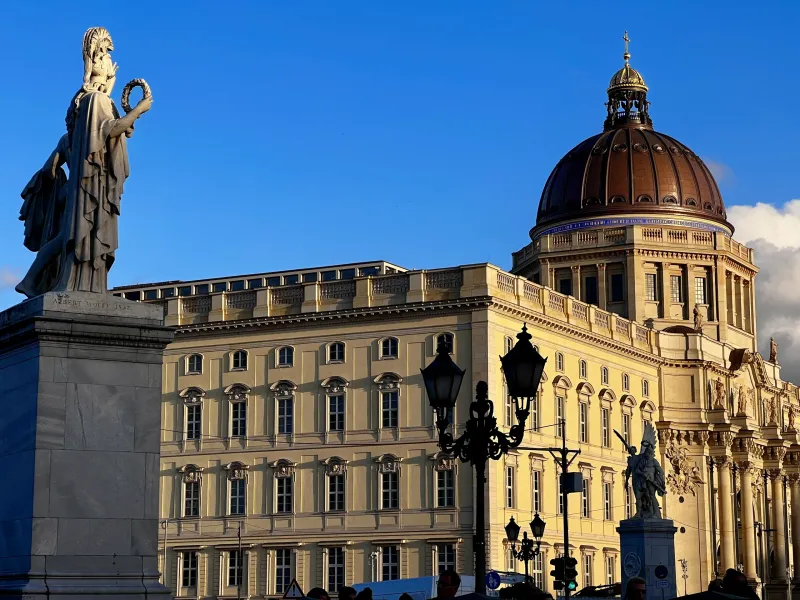A classical stone statue of a female figure holding a wreath, with the dome and facade of the Humboldt Forum in Berlin beh...